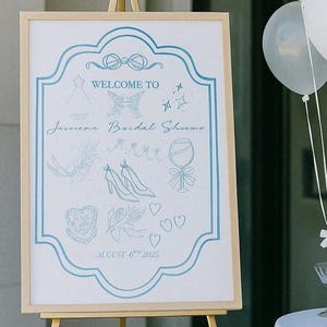 Puede incluir: Un caballete de madera exhibe un letrero de bienvenida con detalles en azul claro. El letrero dice "Welcome to Jessica Bridal Sharma" con ilustraciones decorativas. Globos blancos y copas de martini están dispuestos cerca, sugiriendo un evento nupcial.