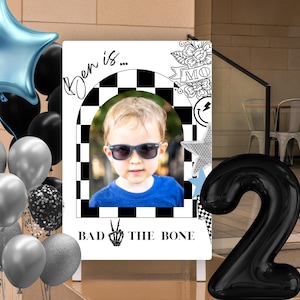 May include: A birthday party decoration featuring a photo of a child wearing sunglasses, with the text "Ben is... BAD THE BONE." The display includes black and silver balloons, a blue star-shaped balloon, and a large black number two balloon.