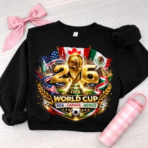 K&ouml;nnte beinhalten: Schwarzes Sweatshirt mit einer goldenen FIFA-Weltmeisterschaftstroph&auml;e und der Zahl 26. Das Design zeigt die Flaggen der USA, Kanadas und Mexikos, zusammen mit dem Text "USA - Canada - Mexico" und Fu&szlig;b&auml;llen.