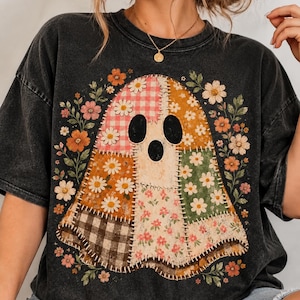 May include: A black acid-wash t-shirt featuring a central graphic of a ghost composed of various patchwork fabric patterns, including gingham, floral, and daisy prints, surrounded by a decorative border of colorful flowers and green leaves.