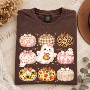 May include: A dark brown t-shirt featuring a graphic print of a central white ghost holding a small pumpkin, surrounded by eight decorative pumpkins with various patterns including daisies, leopard print, plaid, hearts, sunflowers, and autumn leaves.