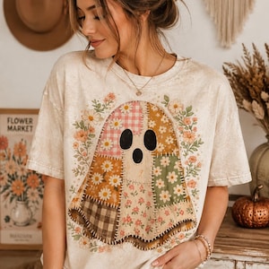 May include: A cream-colored short-sleeve t-shirt featuring a large graphic of a ghost made of various patchwork fabric patterns, including gingham, floral, and plaid prints, surrounded by a wreath of small orange and white flowers.