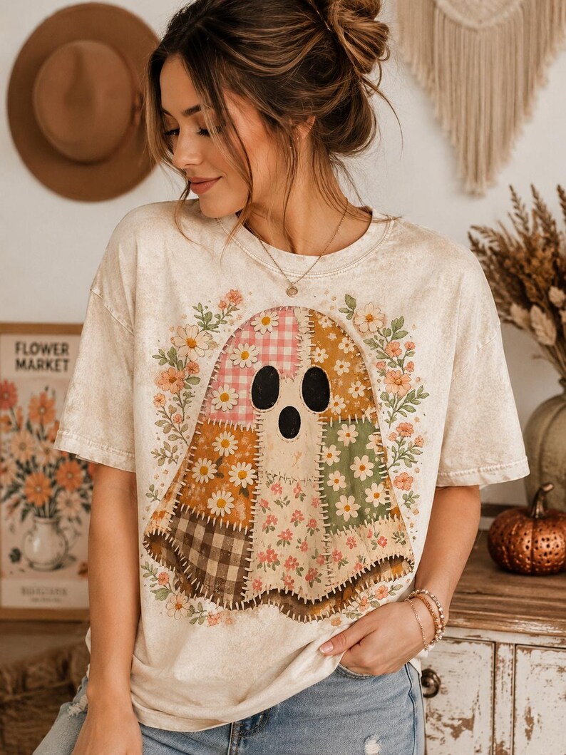 May include: A cream-colored short-sleeve t-shirt featuring a large graphic of a ghost made of various patchwork fabric patterns, including gingham, floral, and plaid prints, surrounded by a wreath of small orange and white flowers.