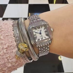 May include: A silver wristwatch with a square face and Roman numeral hour markers. The watch is on a silver link band and is worn with several silver bracelets and a gold-toned bracelet. The watch face is white with the word "GREAY" printed on it.