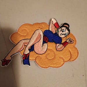 May include: Embroidered patch featuring a cartoon woman in a blue dress and red accents, reclining on an orange cloud. The patch has a white border and is likely designed for application on clothing or accessories.