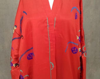 May include: A vibrant red kimono-style jacket with long sleeves. The jacket features embroidered floral and abstract designs in blue, purple, and yellow. The edges are trimmed with a thin green line, and it has matching buttons.