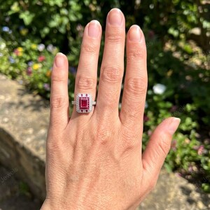 May include: A silver-toned ring featuring a rectangular-cut red gemstone at the center, surrounded by a halo of small white stones. The band is thin and metallic, showcasing a geometric design with a prominent red stone set in a rectangular frame.