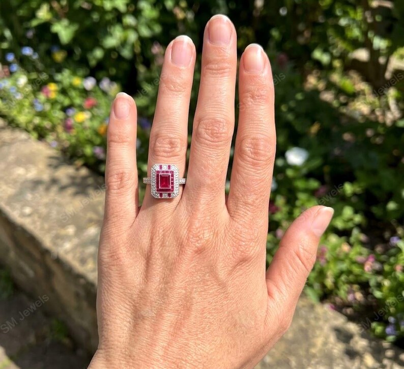 May include: A silver-toned ring featuring a rectangular-cut red gemstone at the center, surrounded by a halo of small white stones. The band is thin and metallic, showcasing a geometric design with a prominent red stone set in a rectangular frame.