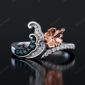 May include: A silver ring featuring a wave-shaped design adorned with small clear white stones and a row of blue stones. A rose gold-colored hibiscus flower sits prominently on one side of the wave, with a band encrusted with clear white stones.