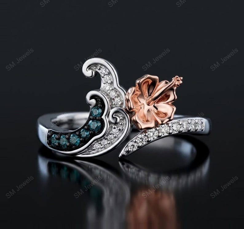 May include: A silver ring featuring a wave-shaped design adorned with small clear white stones and a row of blue stones. A rose gold-colored hibiscus flower sits prominently on one side of the wave, with a band encrusted with clear white stones.