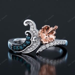 May include: A silver ring featuring a wave-shaped design adorned with small clear stones and a cluster of dark blue stones. A rose gold hibiscus flower sits prominently on one side of the wave, with additional clear stones along the band.