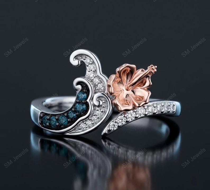 May include: A silver ring featuring a wave-shaped design adorned with small clear stones and a cluster of dark blue stones. A rose gold hibiscus flower sits prominently on one side of the wave, with additional clear stones along the band.
