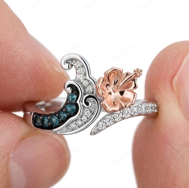 May include: A two-tone ring featuring a silver-colored band with a wave design accented by small blue stones and clear crystals. A rose gold-colored hibiscus flower sits on one side of the wave, with a band of clear crystals along the shank.
