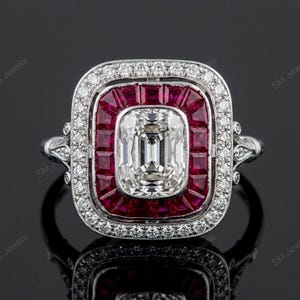May include: A silver-toned ring featuring a central emerald-cut white gemstone surrounded by a rectangular halo of deep red rubies. An outer border of small white diamonds frames the rubies, with additional diamonds set along the metal band.