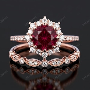 May include: A rose gold engagement ring set with a large, round, deep red gemstone surrounded by smaller white stones. The band is also adorned with small white stones. A matching band sits below.