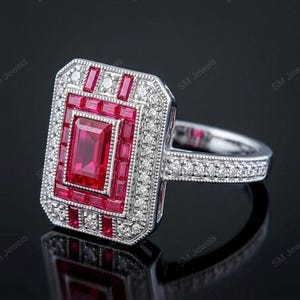 May include: A rectangular silver ring featuring a central emerald-cut red gemstone surrounded by a halo of smaller baguette-cut red stones and a border of round white diamonds. The band is also set with small white diamonds and has milgrain edges.