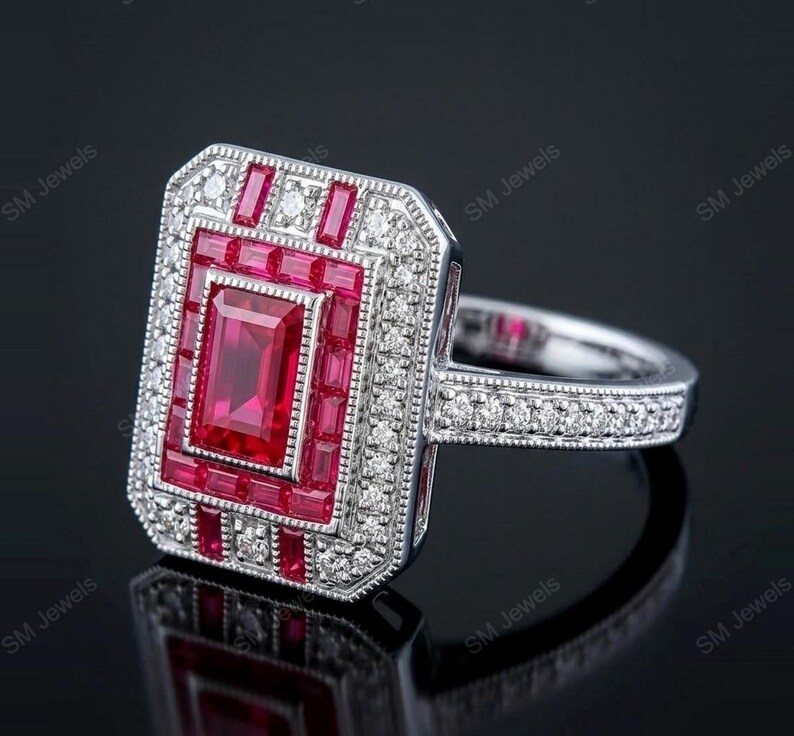 May include: A rectangular silver ring featuring a central emerald-cut red gemstone surrounded by a halo of smaller baguette-cut red stones and a border of round white diamonds. The band is also set with small white diamonds and has milgrain edges.