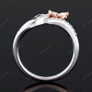 May include: A two-tone ring featuring a polished silver-colored band with a row of small clear stones and a rose gold-colored accent piece at the top. The design includes a curved, split-shank structure with the text SM Jewels visible.