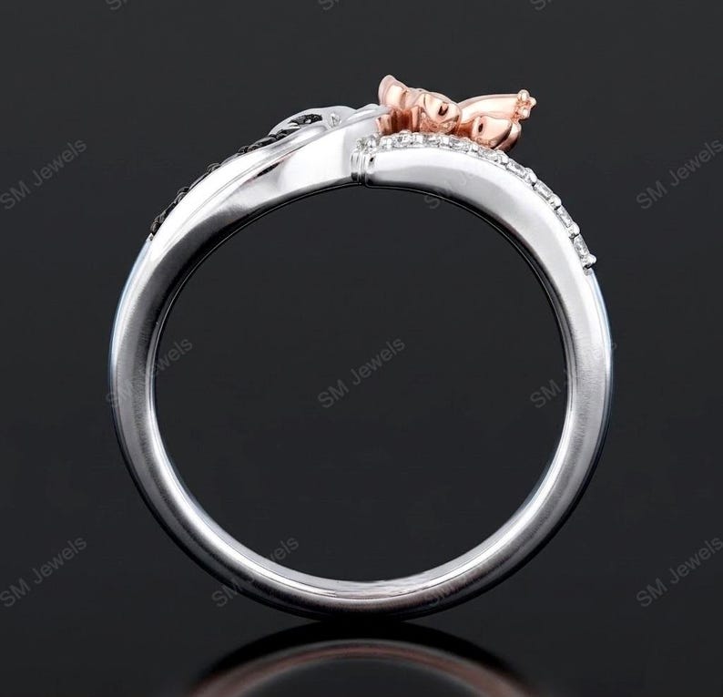 May include: A two-tone ring featuring a polished silver-colored band with a row of small clear stones and a rose gold-colored accent piece at the top. The design includes a curved, split-shank structure with the text SM Jewels visible.