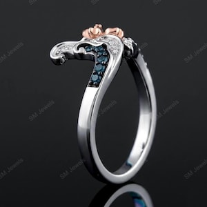 May include: A silver-toned ring featuring a curved design with a rose gold-toned top accent. The band is embellished with a row of small, dark blue stones and a row of clear, white stones set along the side, creating a layered, multi-toned appearance.