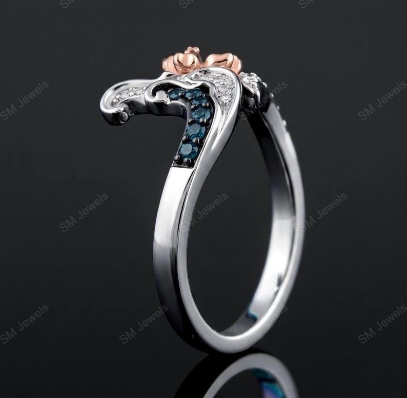 May include: A silver-toned ring featuring a curved design with a rose gold-toned top accent. The band is embellished with a row of small, dark blue stones and a row of clear, white stones set along the side, creating a layered, multi-toned appearance.