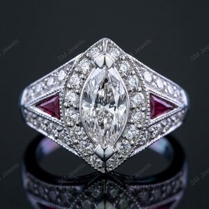 Puede incluir: Un anillo de diamantes con corte marquesa y engaste de oro blanco. El anillo presenta un gran diamante central, rodeado de diamantes más pequeños y dos gemas rojas triangulares. El anillo está sobre un fondo oscuro.