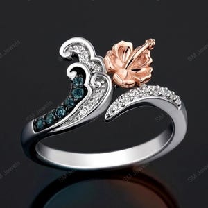 May include: A silver ring featuring a wave-shaped design adorned with small clear and blue stones. A rose gold hibiscus flower sits at the top of the wave. The band is polished silver, and the overall structure creates a curved, open-ended appearance.