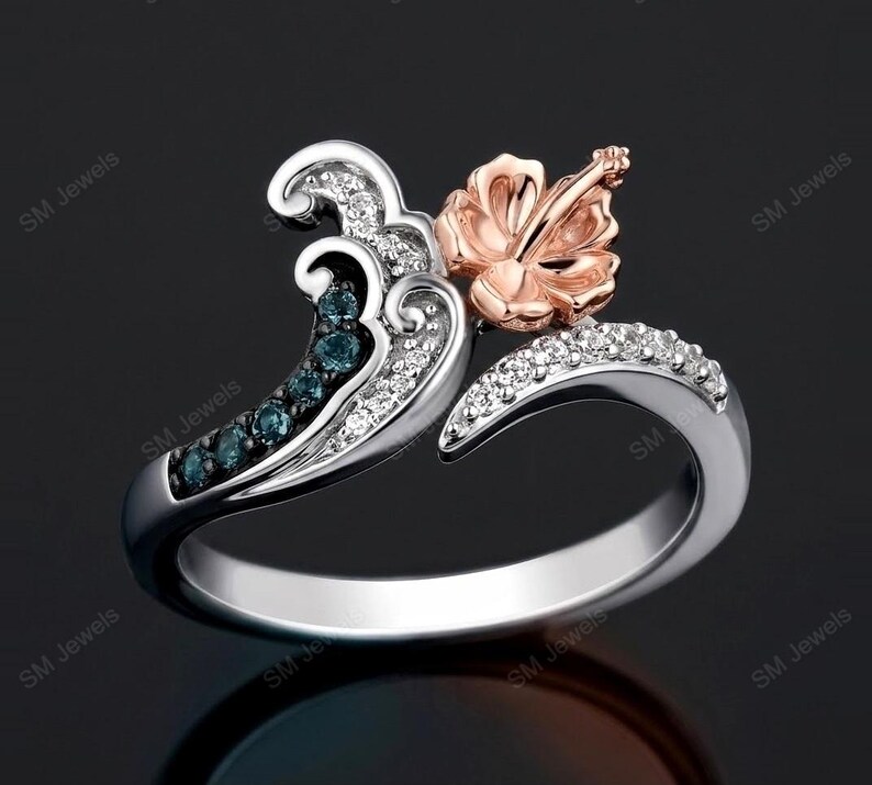May include: A silver ring featuring a wave-shaped design adorned with small clear and blue stones. A rose gold hibiscus flower sits at the top of the wave. The band is polished silver, and the overall structure creates a curved, open-ended appearance.