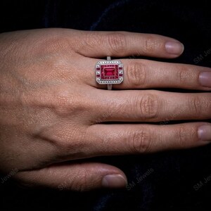 May include: A silver-toned ring featuring a rectangular, deep red center stone surrounded by a border of smaller, clear white stones. The setting has a geometric, stepped design with additional red accents framing the central gemstone.