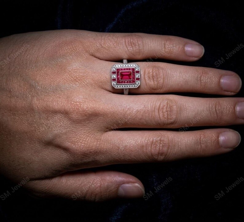 May include: A silver-toned ring featuring a rectangular, deep red center stone surrounded by a border of smaller, clear white stones. The setting has a geometric, stepped design with additional red accents framing the central gemstone.