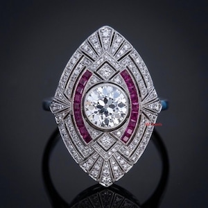 May include: A silver-toned ring featuring a large, round, brilliant-cut center stone surrounded by a marquise-shaped frame. The setting is encrusted with numerous small, clear pavé diamonds and two curved rows of deep red, square-cut rubies.
