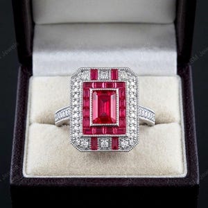 May include: A silver-toned ring featuring a large rectangular center ruby surrounded by a halo of smaller baguette-cut rubies and a border of tiny round white stones. The band is also accented with small white stones along the sides.