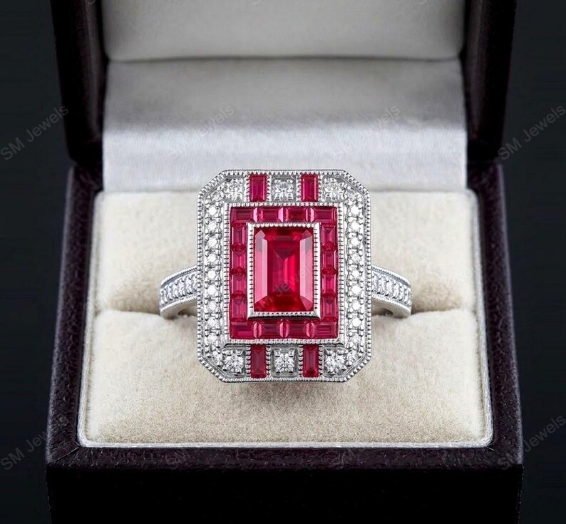 May include: A silver-toned ring featuring a large rectangular center ruby surrounded by a halo of smaller baguette-cut rubies and a border of tiny round white stones. The band is also accented with small white stones along the sides.