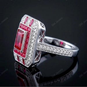 May include: A silver-toned ring featuring a large rectangular center ruby surrounded by a border of smaller baguette-cut rubies and a halo of tiny round white stones. The band is decorated with a row of small white stones and milgrain detailing.