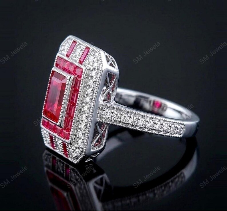 May include: A silver-toned ring featuring a large rectangular center ruby surrounded by a border of smaller baguette-cut rubies and a halo of tiny round white stones. The band is decorated with a row of small white stones and milgrain detailing.
