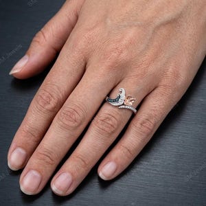 May include: A two-tone ring featuring a silver-colored band with a curved design, accented by small clear stones. One side displays a dark blue, crescent-shaped element, while the other side features a rose gold-colored flower-shaped detail.
