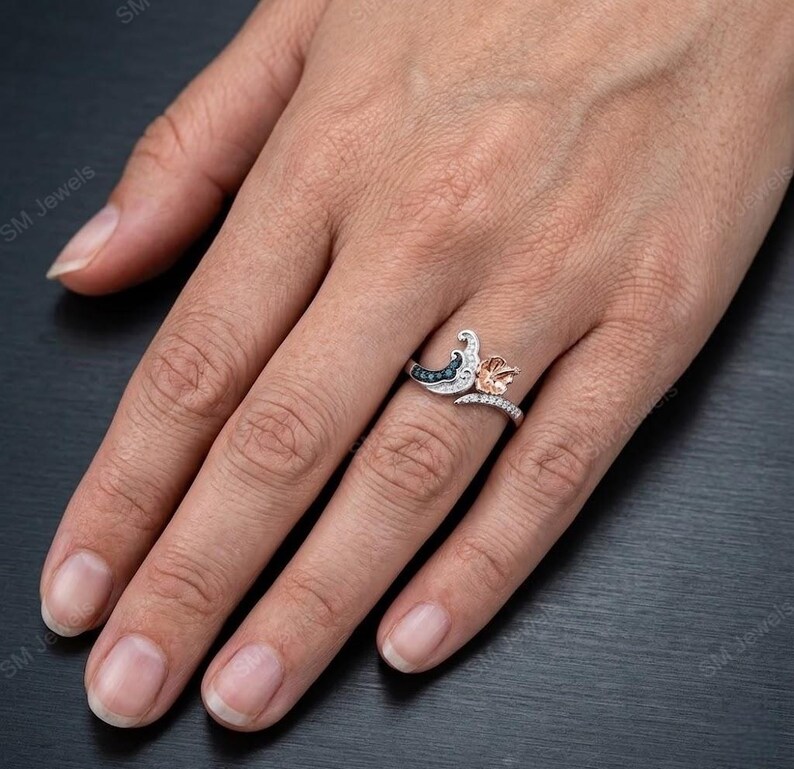 May include: A two-tone ring featuring a silver-colored band with a curved design, accented by small clear stones. One side displays a dark blue, crescent-shaped element, while the other side features a rose gold-colored flower-shaped detail.