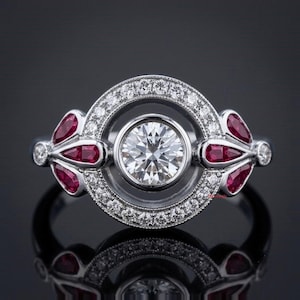 Puede incluir: Un anillo de tono plateado con un diamante central de talla brillante redonda engastado en un halo circular de diamantes más pequeños. El diseño incluye tres rubíes rojos en forma de pera a cada lado, acentuados por pequeños diamantes redondos.