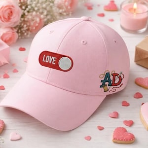 May include: A light pink baseball cap with the word "LOVE" on a red rectangle and a white circle. The cap also features an embroidered "AD" design with a needle and thread. The cap is surrounded by heart-shaped cookies and a lit candle.