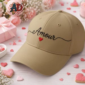 Op de afbeelding: Beige baseballpet met het woord "Amour" in zwart geborduurd, met een klein rood hart. De pet heeft een gebogen klep en een klassiek zes-panelen ontwerp. De achtergrond bevat roze harten en bloemen.