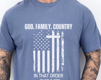 May include: Ice blue t-shirt with a distressed American flag design and a cross. The text reads "GOD. FAMILY. COUNTRY IN THAT ORDER - CHARLIE KIRK". The shirt is a short-sleeved crew neck.