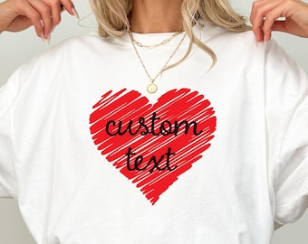 May include: White t-shirt featuring a large red heart with the words "custom text" in a handwritten style. The heart design is filled with red scribbled lines. The shirt is a casual style, suitable for everyday wear.