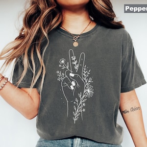May include: Dark gray t-shirt featuring a white hand making a peace sign with floral accents. The design includes detailed white line art of flowers and leaves. The shirt is paired with blue jeans and white slip-on shoes. The word "Pepper" is in the top right corner.