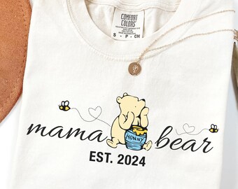 May include: Ivory-colored t-shirt featuring a Winnie the Pooh graphic with the words "mama bear" and "EST. 2024" in black script. The design includes bees and hearts. A gold necklace is draped on the shirt.