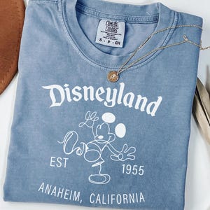 Puede incluir: Camiseta azul hielo con el logotipo de Disneyland y un gr&aacute;fico de Mickey Mouse. La camiseta incluye el texto "EST 1955 ANAHEIM, CALIFORNIA". Un collar dorado est&aacute; sobre la camiseta.