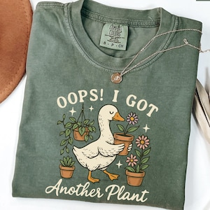 May include: Moss green t-shirt with the text "Oops! I Got Another Plant" and a cartoon duck holding a potted flower. The shirt has a crew neck and short sleeves. A silver necklace is draped across the shirt.