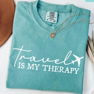 May include: Seafoam green t-shirt with the text "Travel is my Therapy" in white script and a white airplane graphic. A gold necklace with a pendant is on the shirt. A brown hat and dried plant are in the background.