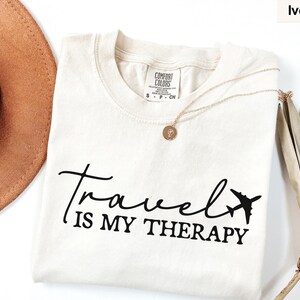 May include: Ivory t-shirt featuring the text "Travel is my Therapy" in black script and a small airplane graphic. A gold necklace with a pendant is on the shirt. A brown hat and dried plant are in the background.
