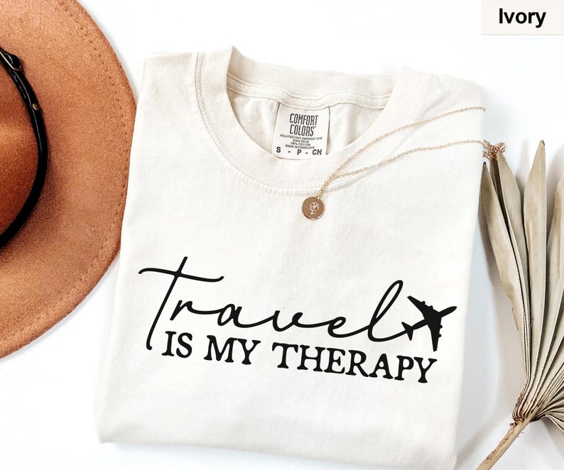 May include: Ivory t-shirt featuring the text "Travel is my Therapy" in black script and a small airplane graphic. A gold necklace with a pendant is on the shirt. A brown hat and dried plant are in the background.