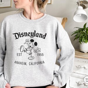 Puede incluir: Una sudadera gris claro con el logotipo de Disneyland en negro, con Mickey Mouse. El texto incluye "EST 1955" y "ANAHEIM, CALIFORNIA". La sudadera es de color gris deportivo.